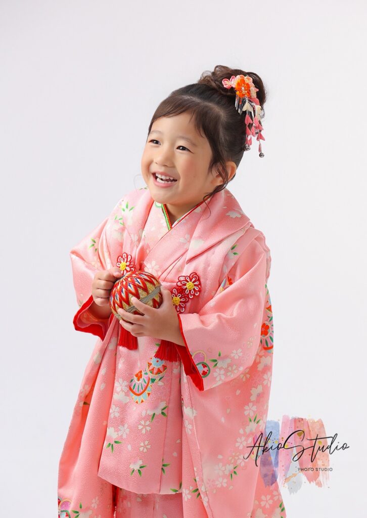 鮮やかなピンク色の着物を身にまとった可愛らしい姿。華やかな髪飾りと、手に持った伝統的な手毬（てまり）が、日本の文化の美しさを引き立てています。