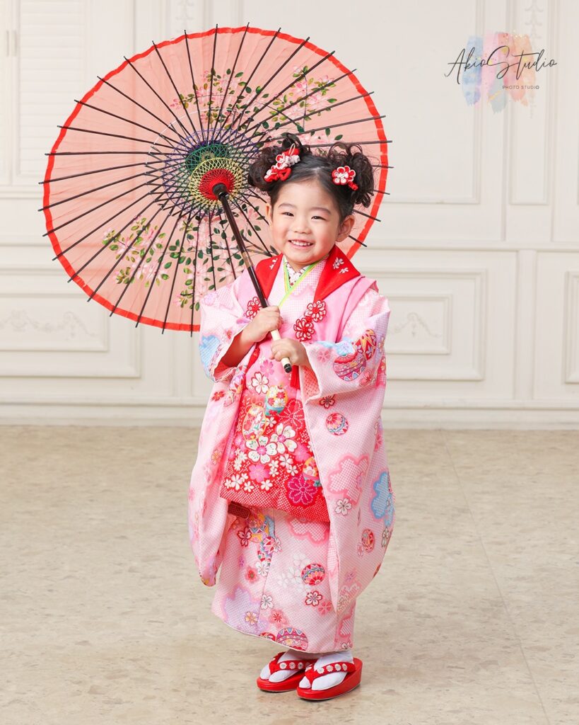 こちらのお写真は、華やかなピンク色の着物を着たお子様。和傘を持って自然な笑顔を見せてくださいました。髪飾りの赤い花がキュートなアクセントになっていて、とても可愛いですね。